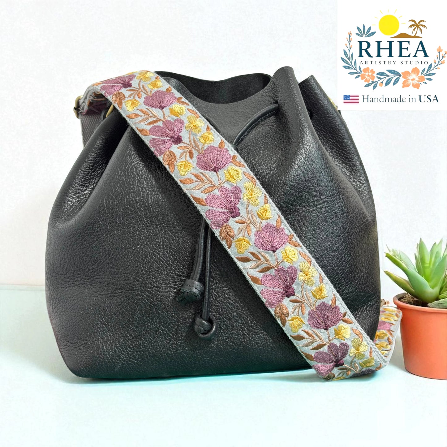 Black leather bucket bag with a floral leather purse strap on a white background, featuring the RHEA brand.