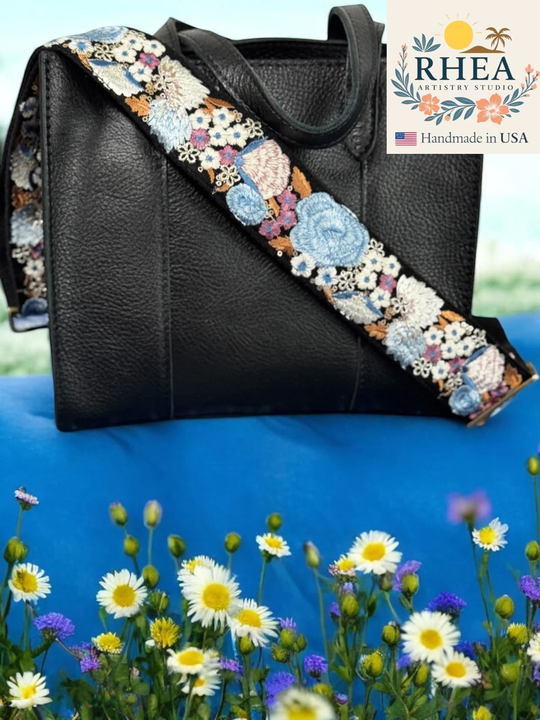 Black handbag with a floral purse strap in a field of flowers, featuring the RHEA Artistry Studio logo.
