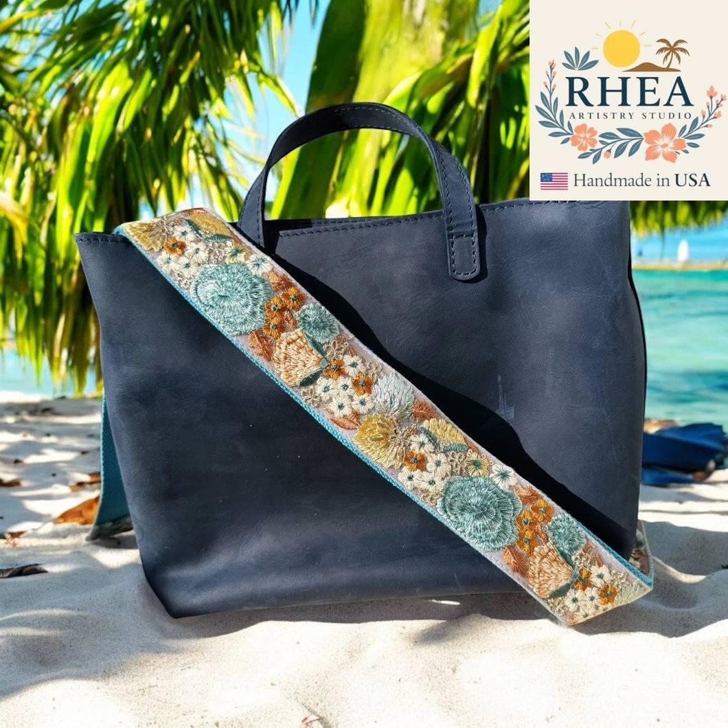 Navy blue tote bag with floral purse strap on a sandy beach, palm trees in the background, RHEA Artistry Studios logo.