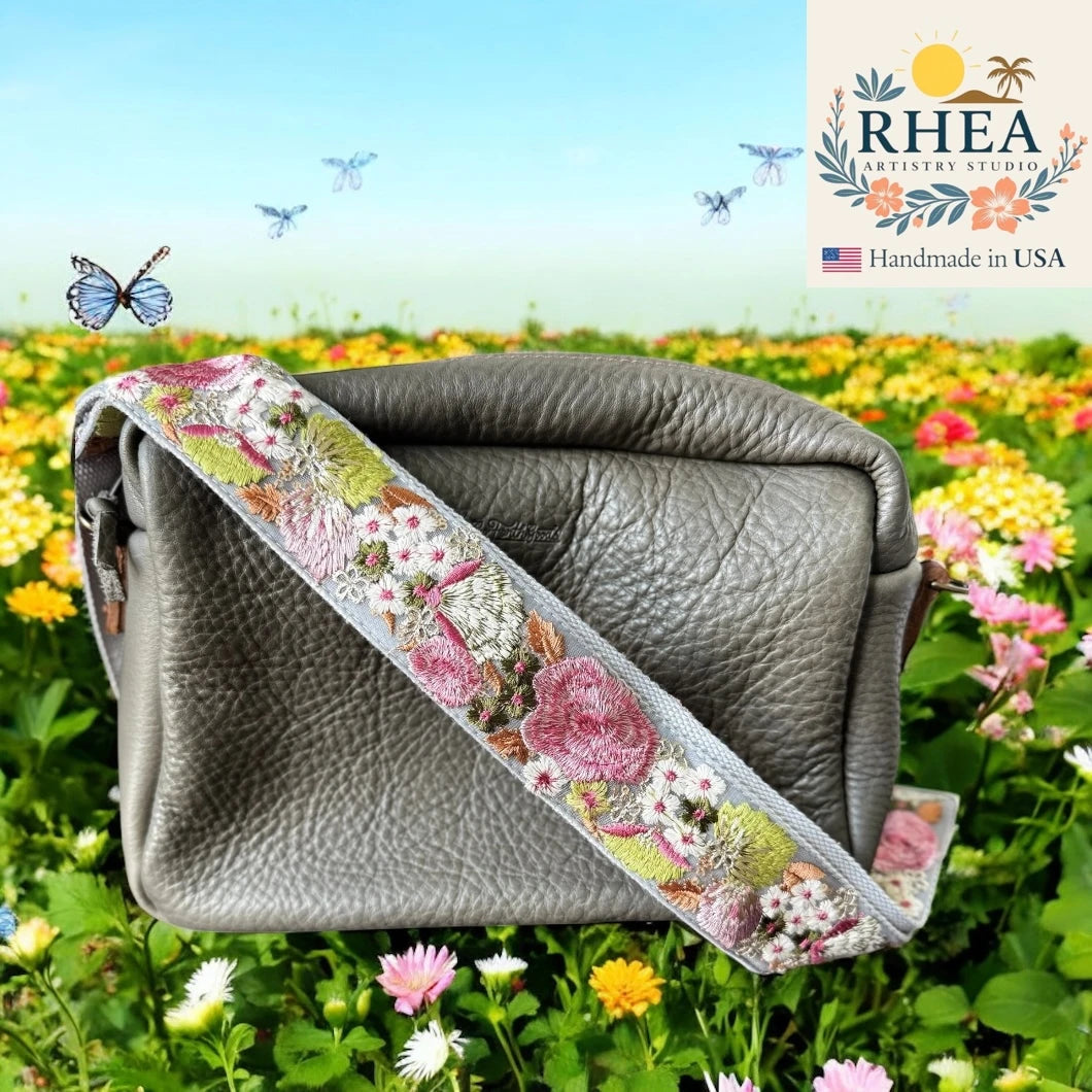 Gray leather bag with floral purse strap against a colorful flower field background, featuring the RHEA Artistry Studios logo.