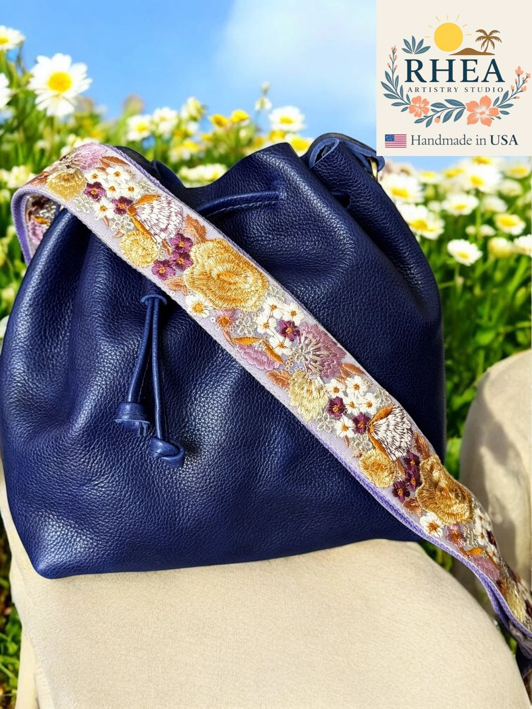 Navy blue leather bag with a floral purse strap in a field of flowers, featuring the RHEA brand.