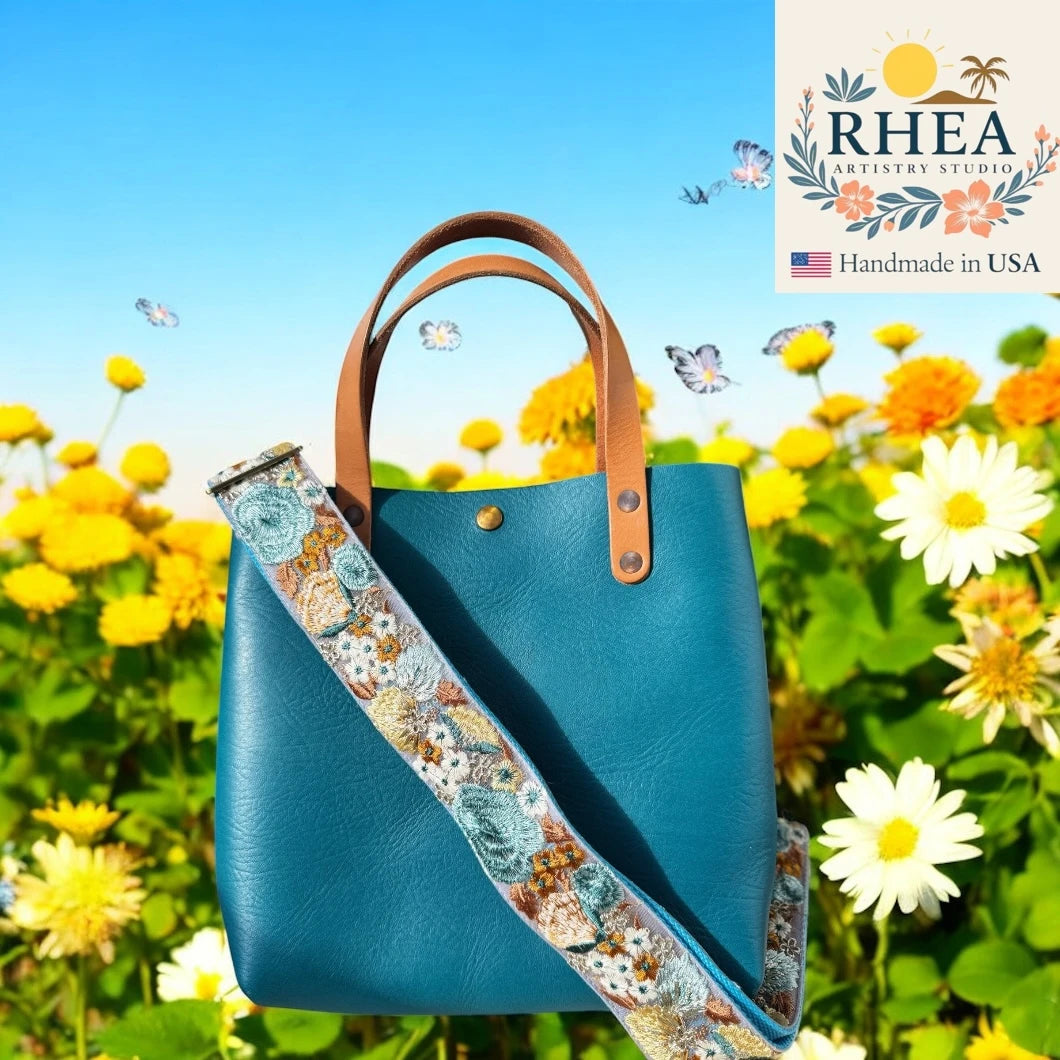 Blue handbag with a floral purse strap in a field of flowers, featuring the RHEA brand.
