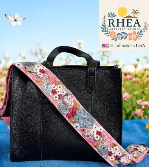 Black handbag with a floral purse strap on a blue surface with flowers in the background, featuring RHEA Artistry Studio branding.