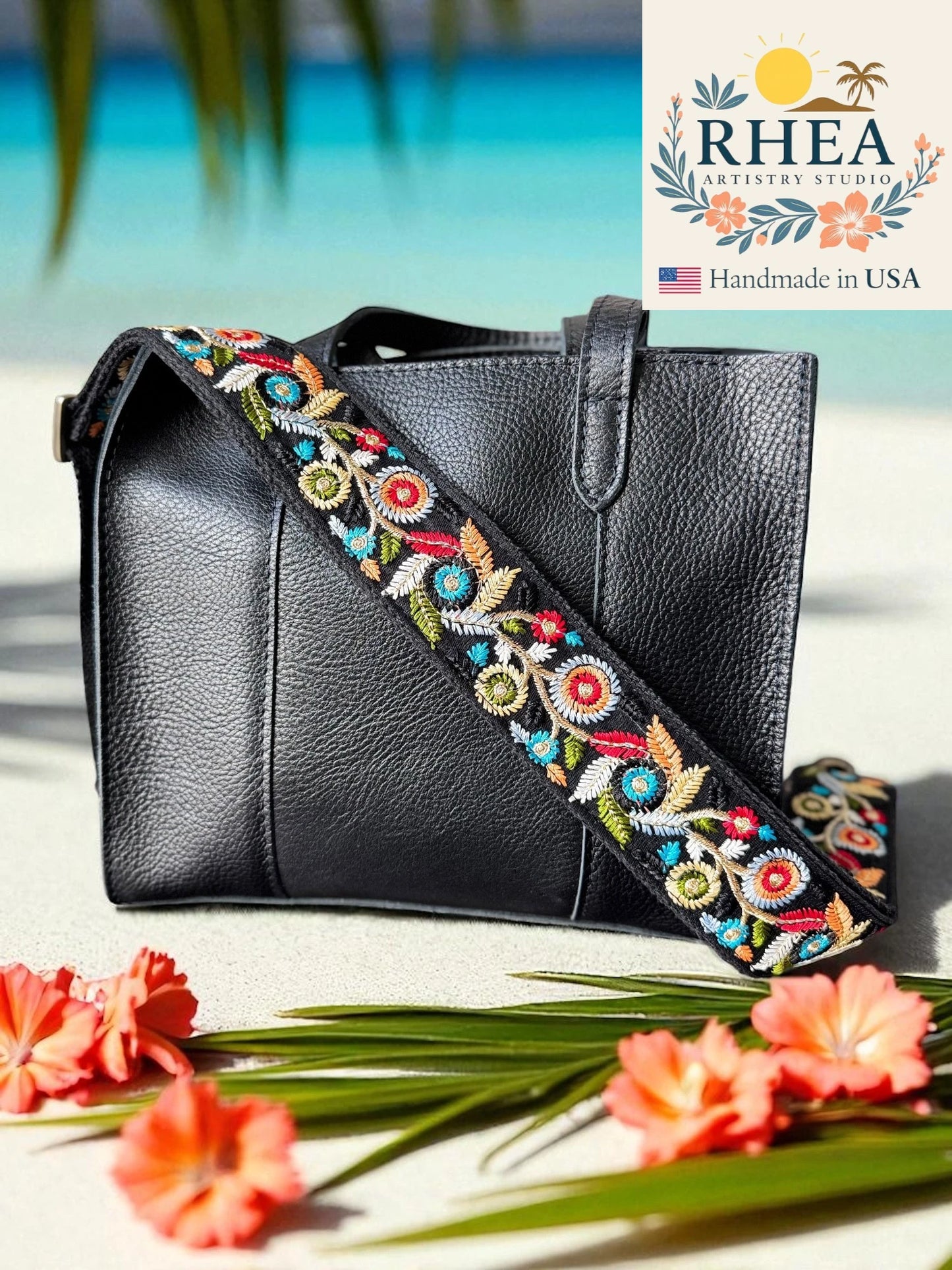 Black handbag with colorful floral strap on a beach setting, featuring the RHEA brand.