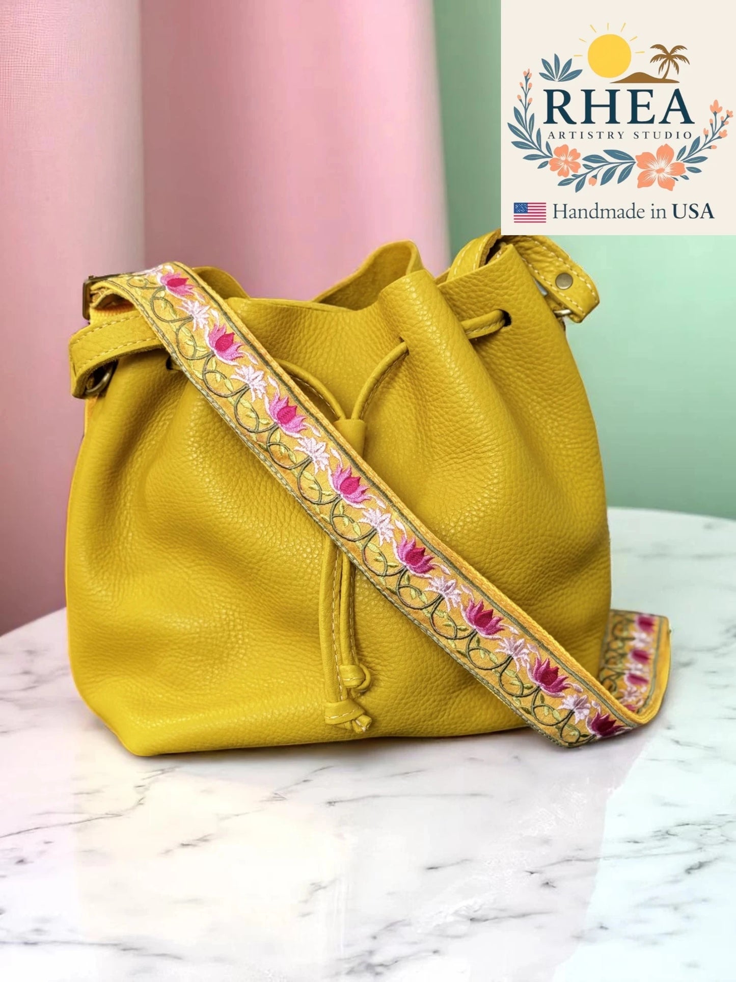 Yellow handbag with floral purse strap on a marble surface, RHEA brand visible.