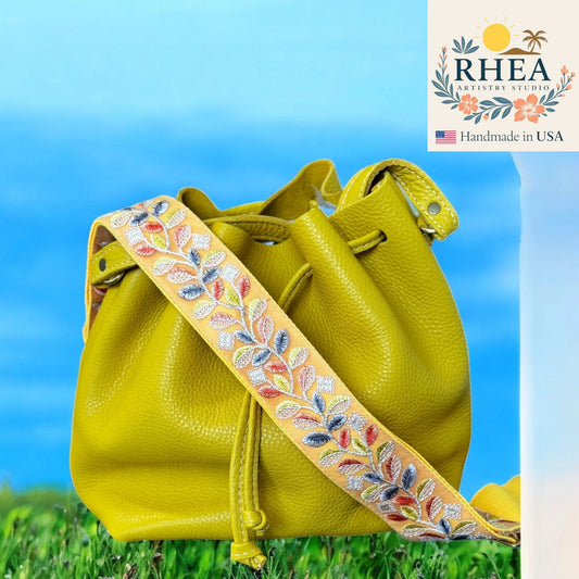 Yellow leather bag with floral leather purse strap against a blue sky and grass background, featuring RHEA Artistry Studio logo.