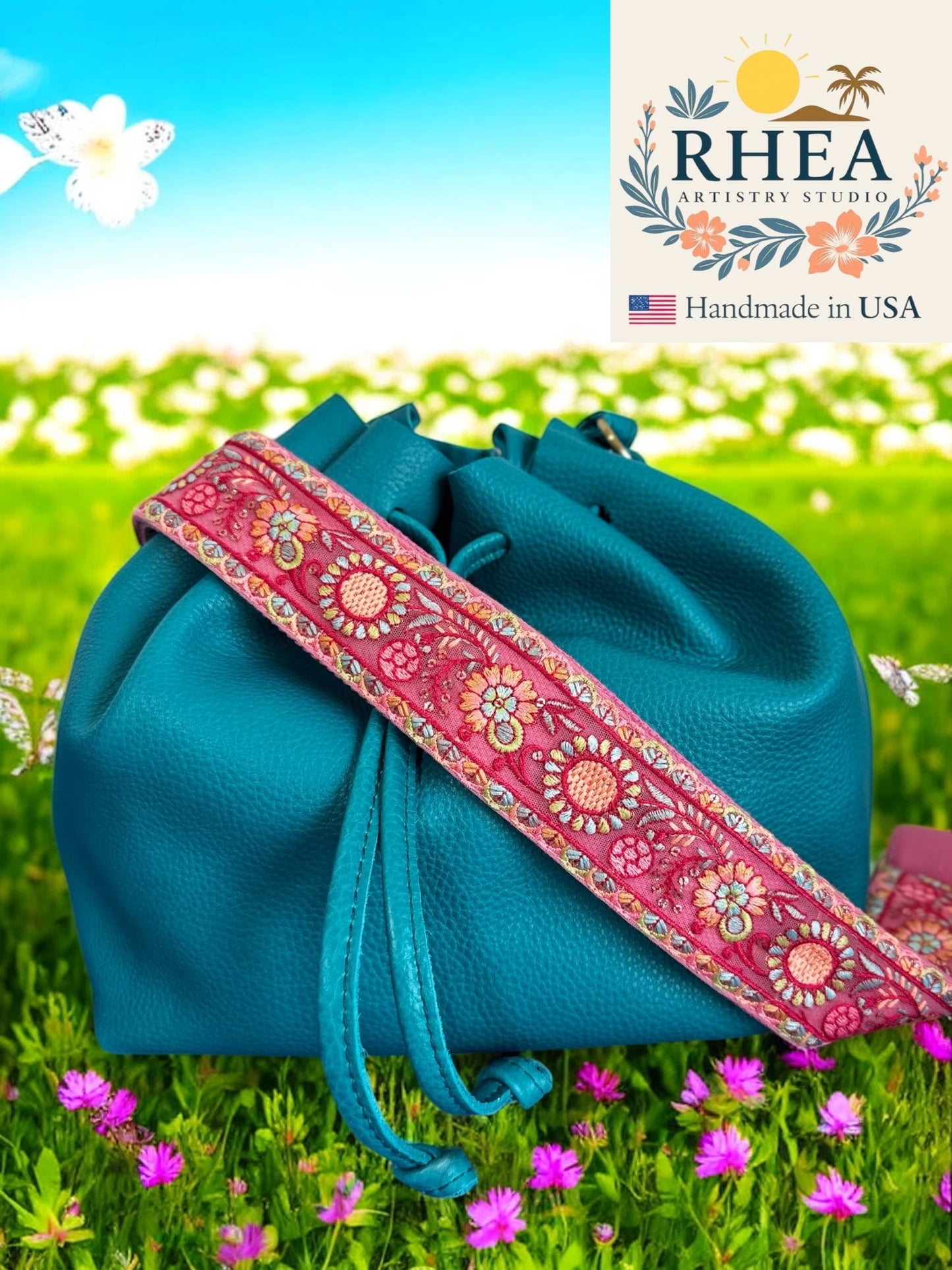Blue bag with a pink floral purse strap on grass with flowers, Rhea Artistry Studio logo.