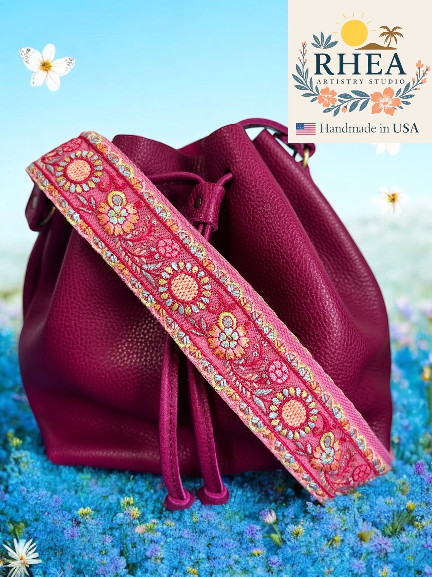 Pink leather bag with a floral purse strap on a blue floral background, featuring the RHEA Antiquities Studio logo.