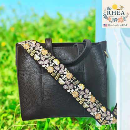 Black handbag with an embroidered strap against a green outdoor background, featuring the RHEA brand.
