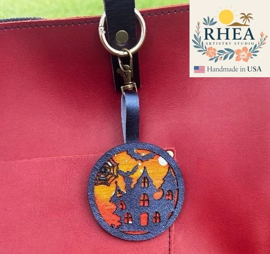 Decorative leather zipper pull charm with Halloween design on a red surface, featuring the RHEA Artistry Studio logo.