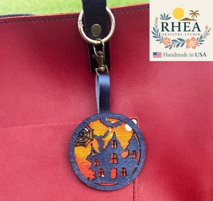 Decorative leather zipper pull charm with Halloween design on a red surface, featuring the RHEA Artistry Studio logo.