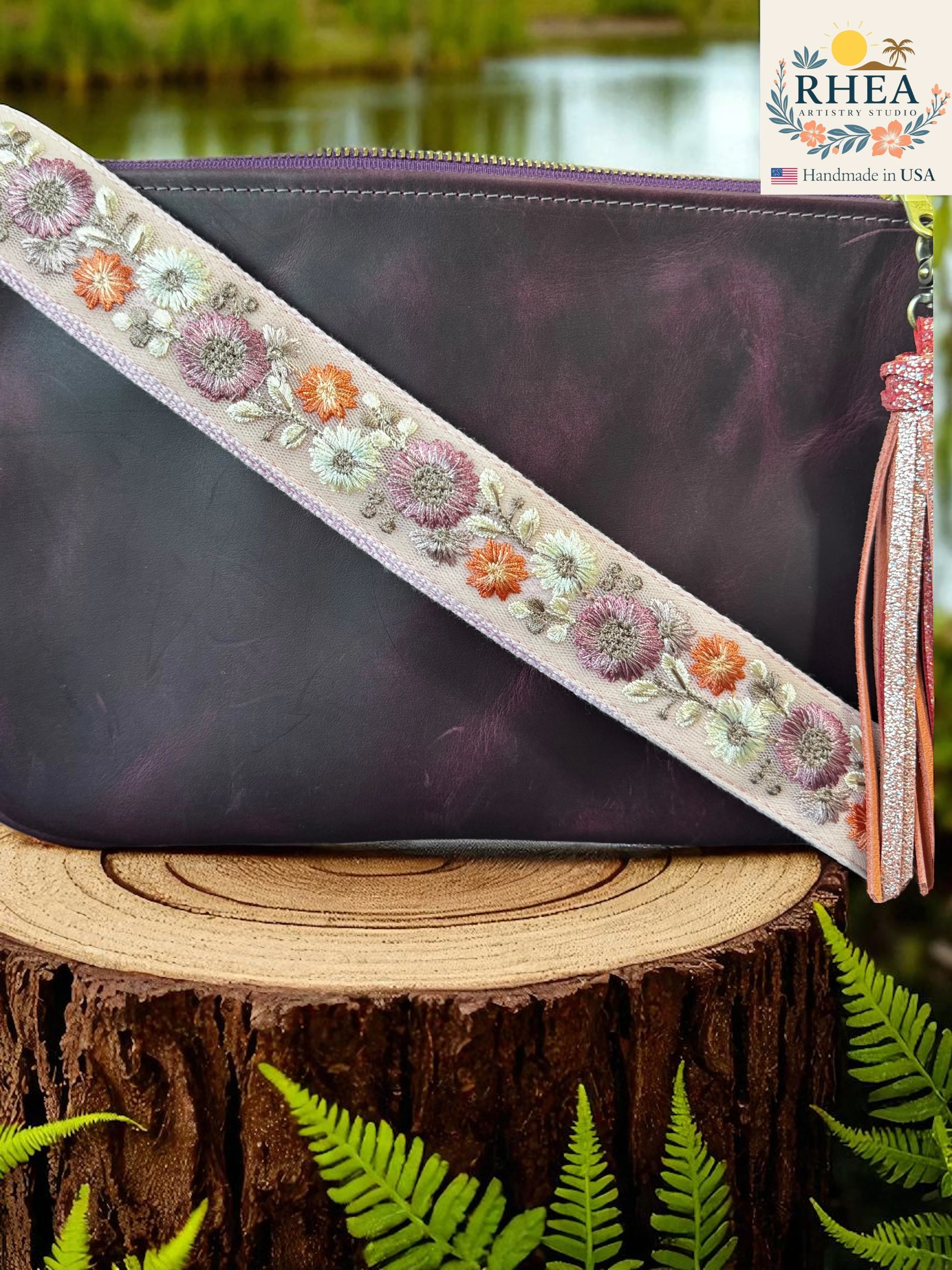 Purple clutch bag with floral strap on a wooden stump with ferns and blurred water background