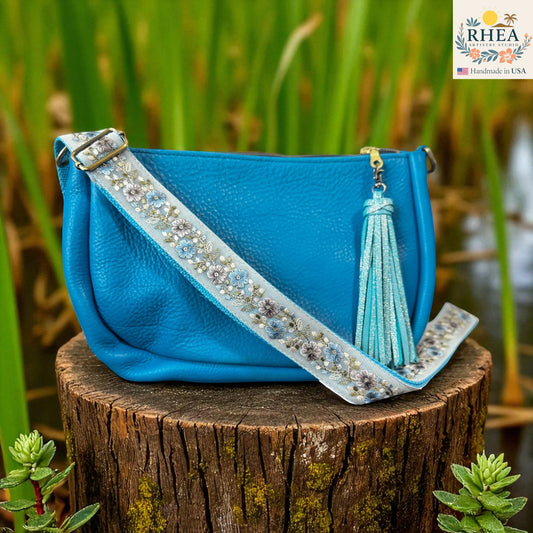 Blue handbag with decorative  artisan strap and tassel on a wooden stump with greenery in the background, featuring the RHEA brand