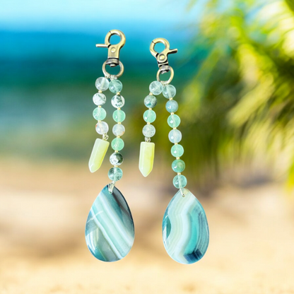 Teal and green beaded earrings with teardrop-shaped stones on a blurred beach background