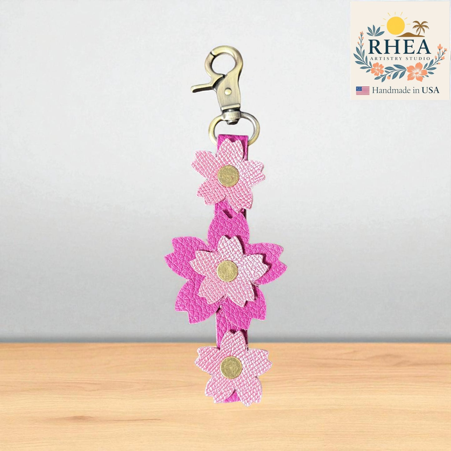 Pink flower-shaped leather purse charm with a clip on a wooden surface, branded 'RHEA'.