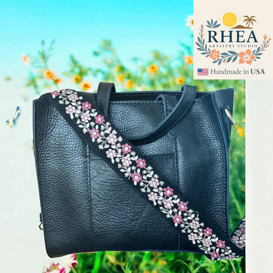 Black handbag with floral strap against a blurred natural background, featuring the RHEA Artisan Studio logo.