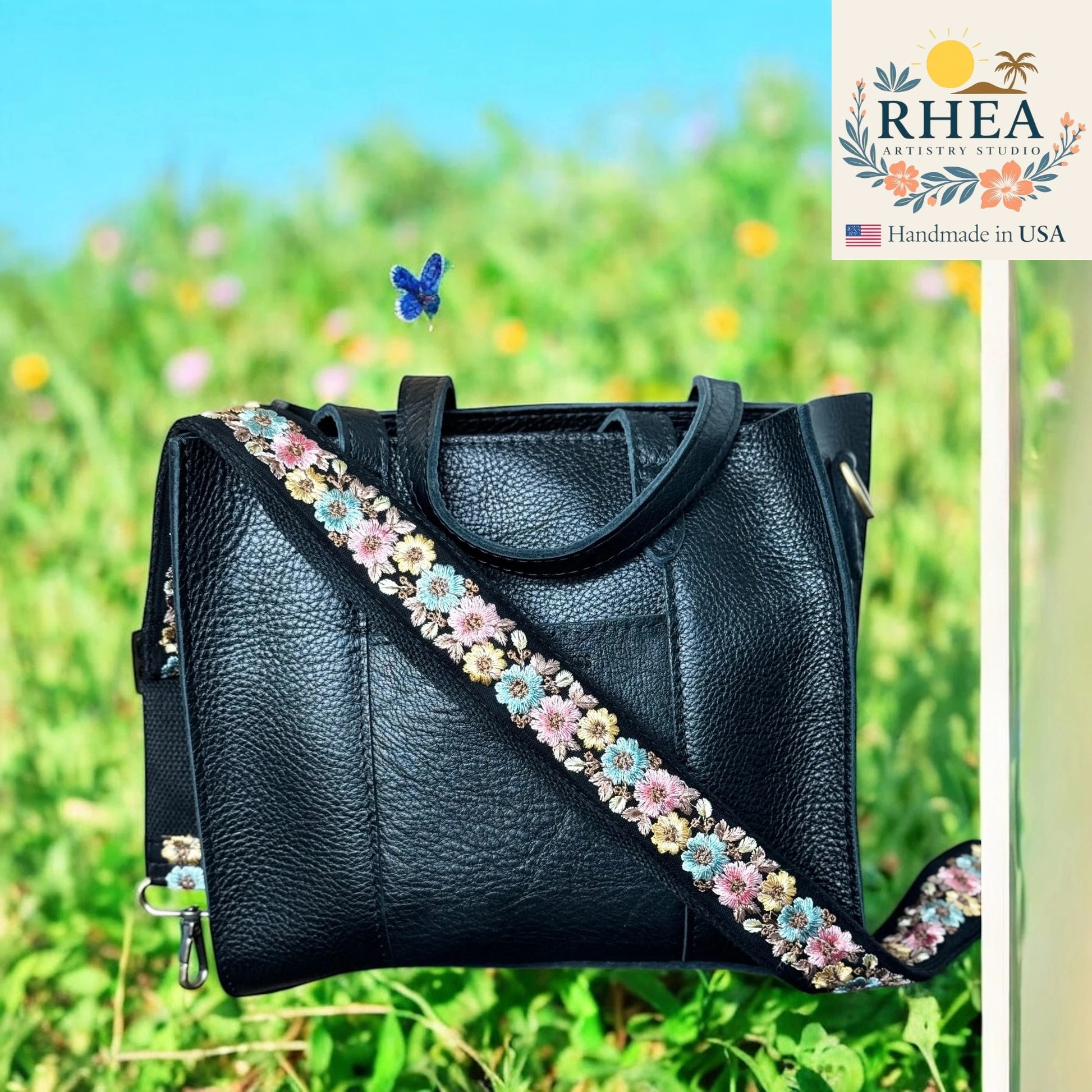 Black leather handbag with floral strap in a grassy field, featuring the RHEA brand.