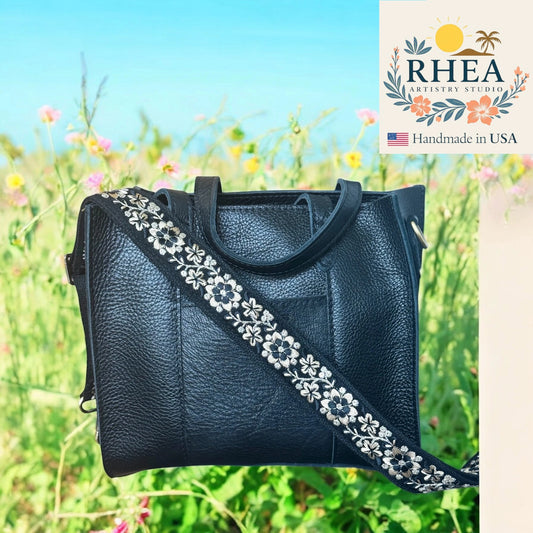 Black leather handbag with floral strap against a natural background, featuring the RHEA Artesian Studio logo.