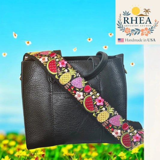 Black handbag with a colorful floral and pineapple patterned strap against a blue sky and green field background. RHEA Artisan Studio logo is visible.