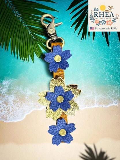 Floral leather purse charm with blue and gold flowers on a beach background, featuring the RHEA brand.
