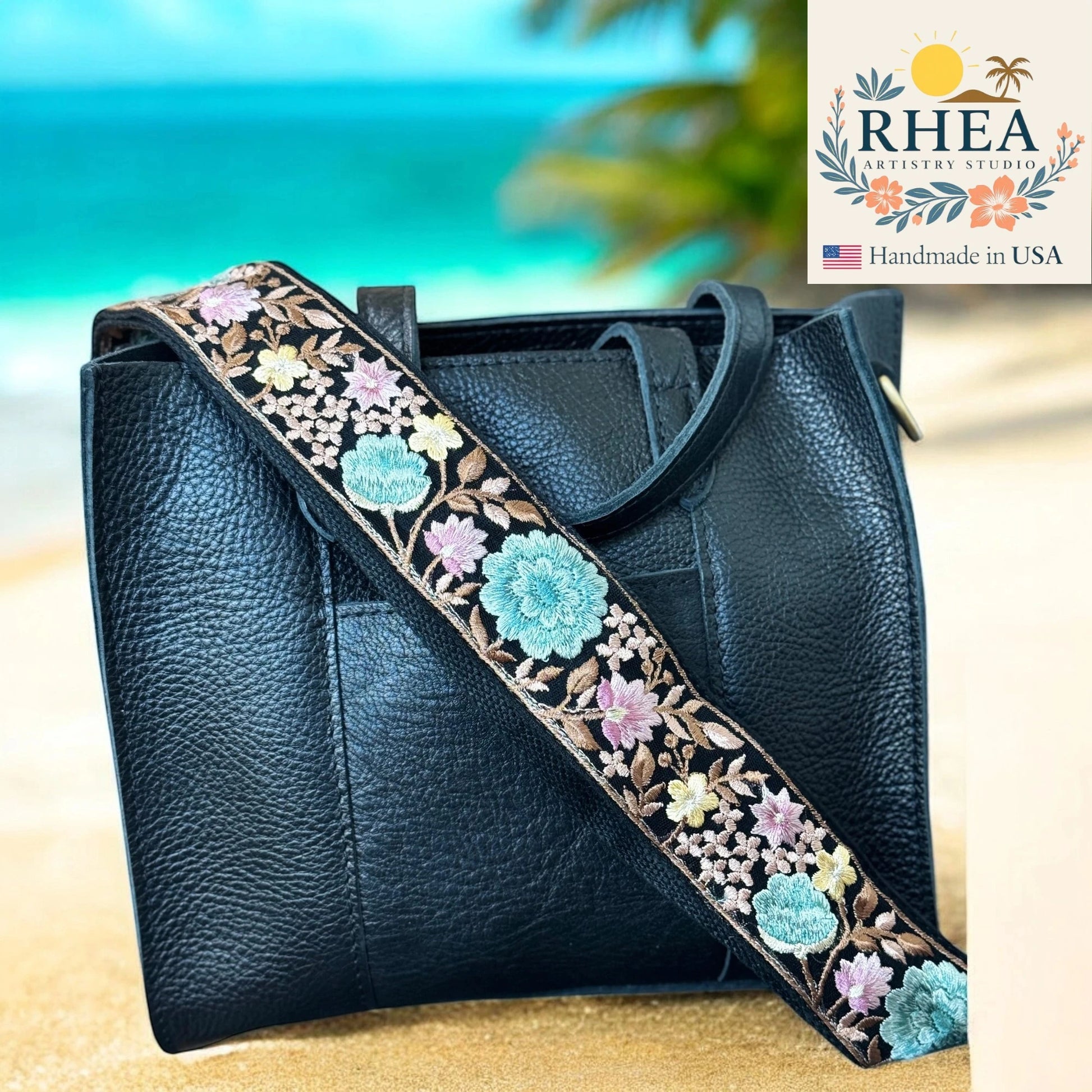 Black leather handbag with floral bag strap on a beach background, featuring RHEA Artistry Studio logo.
