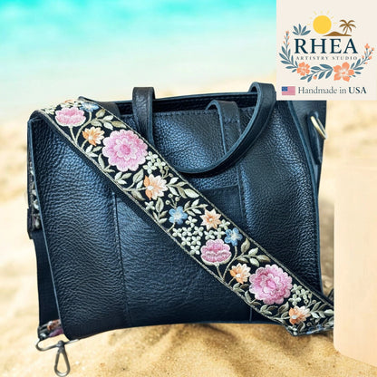 Black leather handbag with floral purse strap on a beach background, featuring the RHEA Artistry Studio logo.