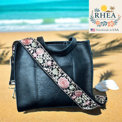Black leather handbag with floral purse strap on a sandy beach, featuring RHEA Artistry Studio logo.