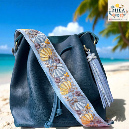 Blue handbag with an embroidered  bag strap on a beach, featuring the RHEEA brand.