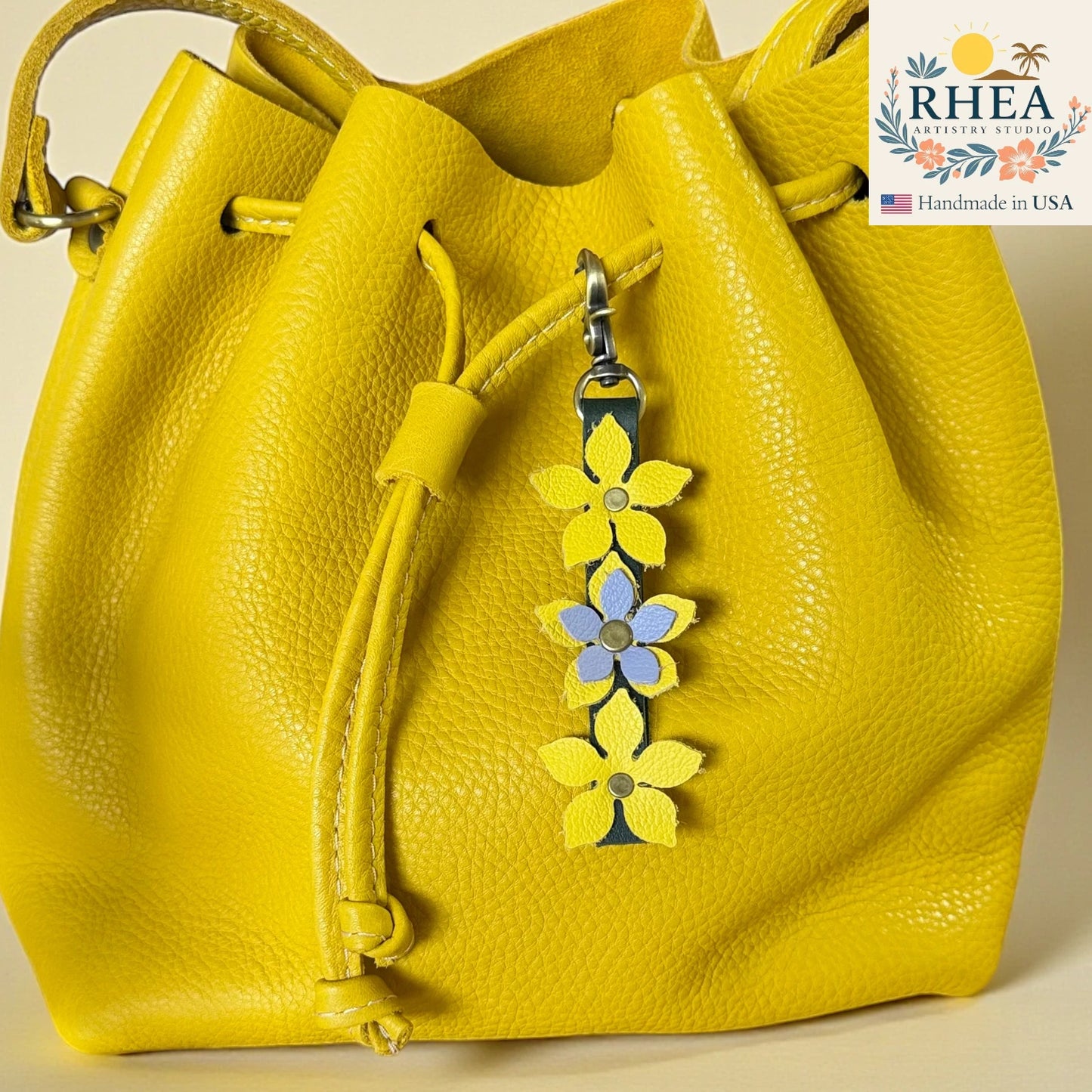 Yellow leather handbag with floral charm and RHEA Artisan Studio logo.