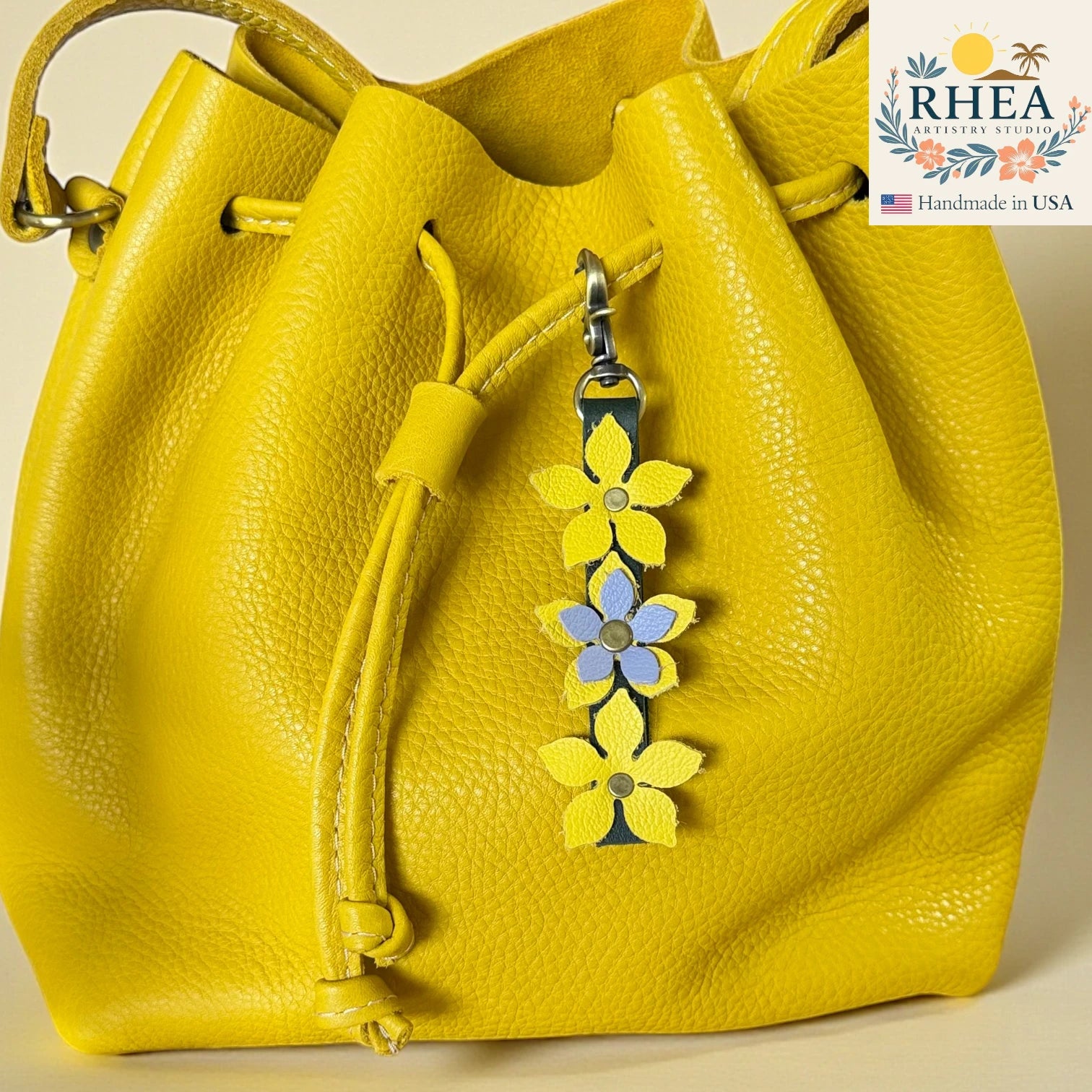 Yellow leather handbag with floral charm and RHEA Artisan Studio logo.