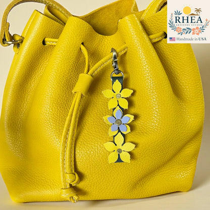 Yellow leather handbag with floral charm and RHEA Artisan Studio logo.