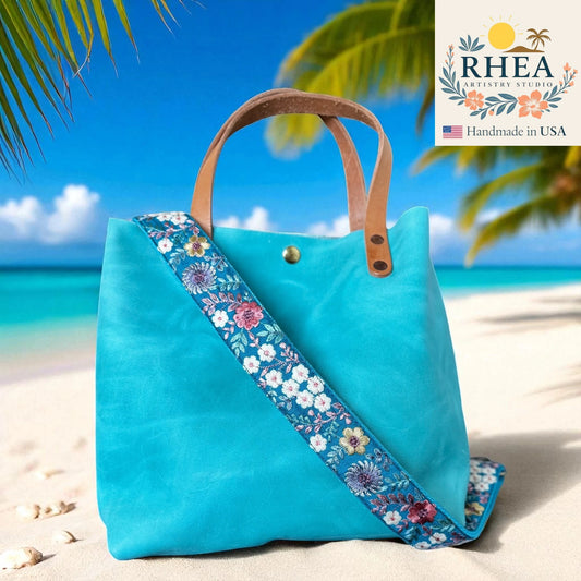 Blue tote bag with floral strap on a beach, featuring RHEA Artistry Studio logo.