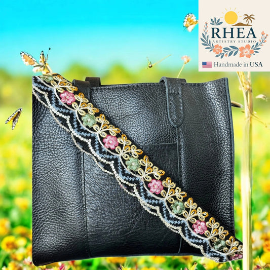 Black leather handbag with decorative strap against a blurred natural background, featuring the Rhea Artistry Studio logo.