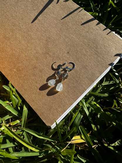 A pair of pale blue silver drop earrings with a cathedral-like design, displayed on a brown paper surface, with blurred greenery in the background.
