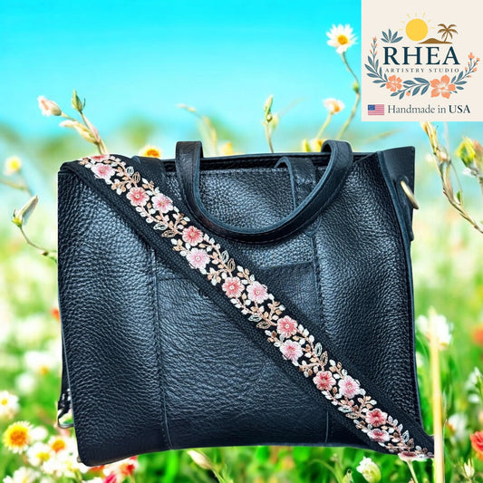 Black leather handbag with floral purse strap against a natural background, featuring the RHEA brand.