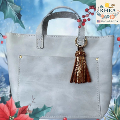 Gray handbag with a brown tassel keychain on a decorative background with holly leaves and berries.