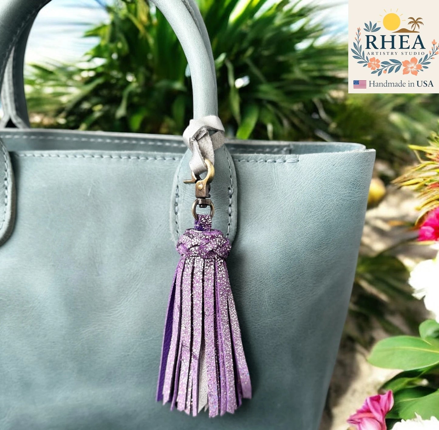Gray handbag with a purple tassel keychain against a natural background, featuring the RHEA Artistry Studio logo.