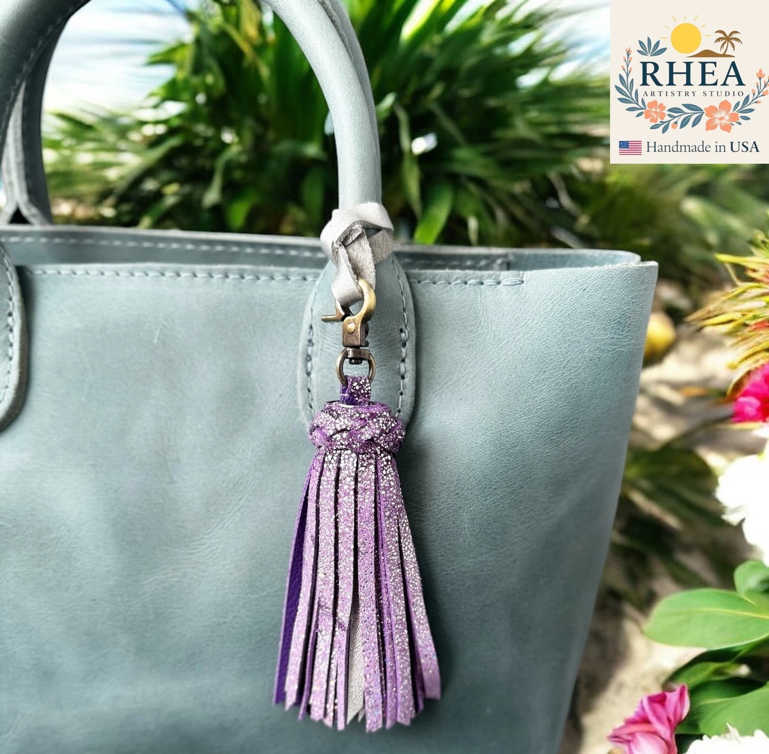 Gray handbag with a purple tassel keychain against a natural background, featuring the RHEA Artistry Studio logo.
