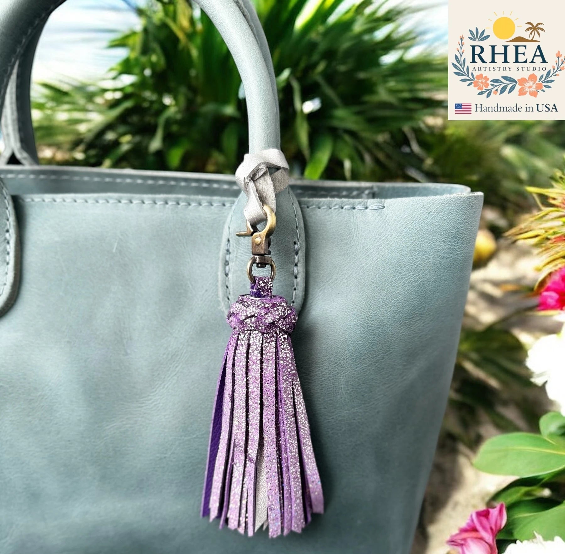 Gray handbag with a purple tassel keychain against a natural background, featuring the RHEA Artistry Studio logo.