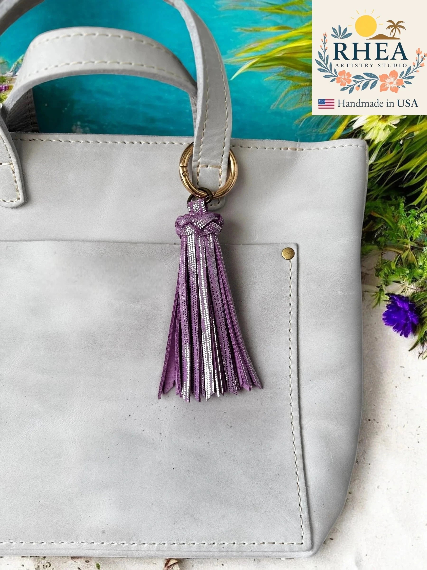 White handbag with a lilac tassel keychain on a blue background, featuring the RHEA brand.