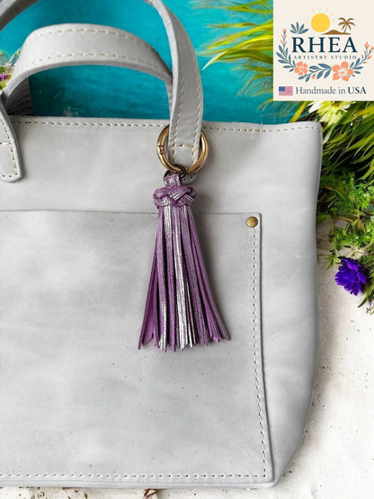 White handbag with a lilac tassel keychain on a blue background, featuring the RHEA brand.