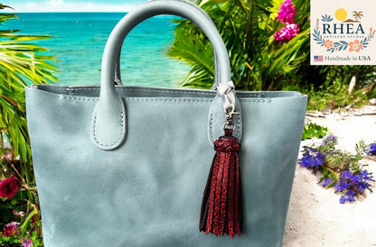 Light blue handbag with a red tassel on a beach background, featuring the RHEA brand.