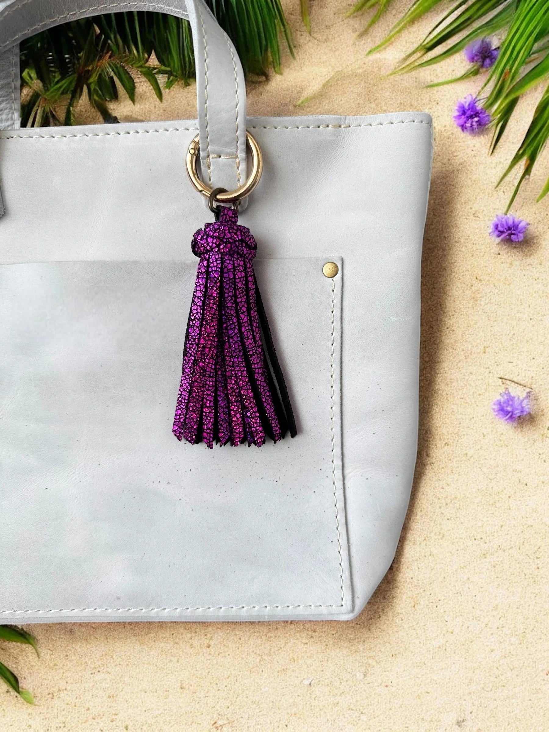 White handbag with a purple tassel keychain on a sandy surface with greenery.