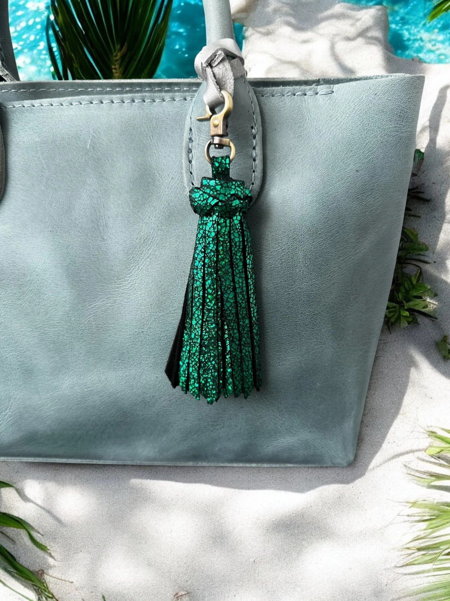 Light blue handbag with a green tassel on a white surface with greenery.