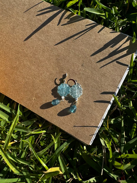 A pair of Daisy Blue Silver Drop Earrings with a blue gemstone, displayed on a brown cardboard surface outdoors.