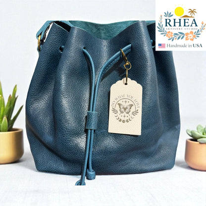 Blue leather bag with a tag on a light background, featuring the RHEA brand.
