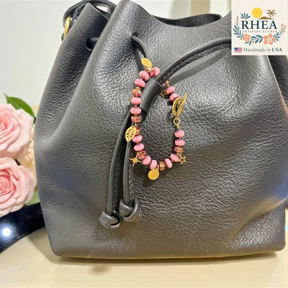 Gray leather handbag with a rose beaded charm, flowers in the background, and RHEA branding.