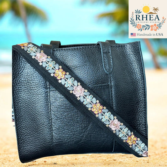Black leather handbag with floral strap on a blurred beach background, featuring the RHEA brand.