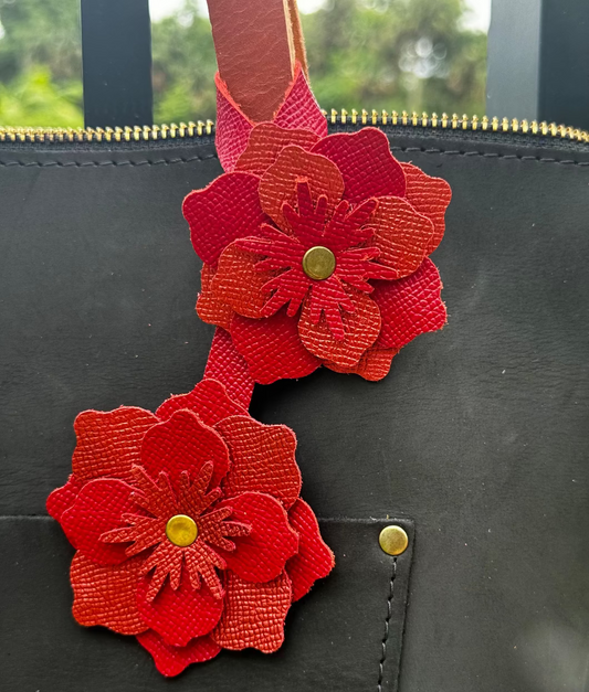 A red and brick red leather flower accessory with a textured surface and brass rivets, designed to attach to a bag or garment.
