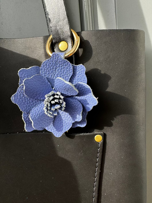 A leather flower accessory with a blue and white color scheme, attached to a black leather-bound notebook with a yellow clasp.